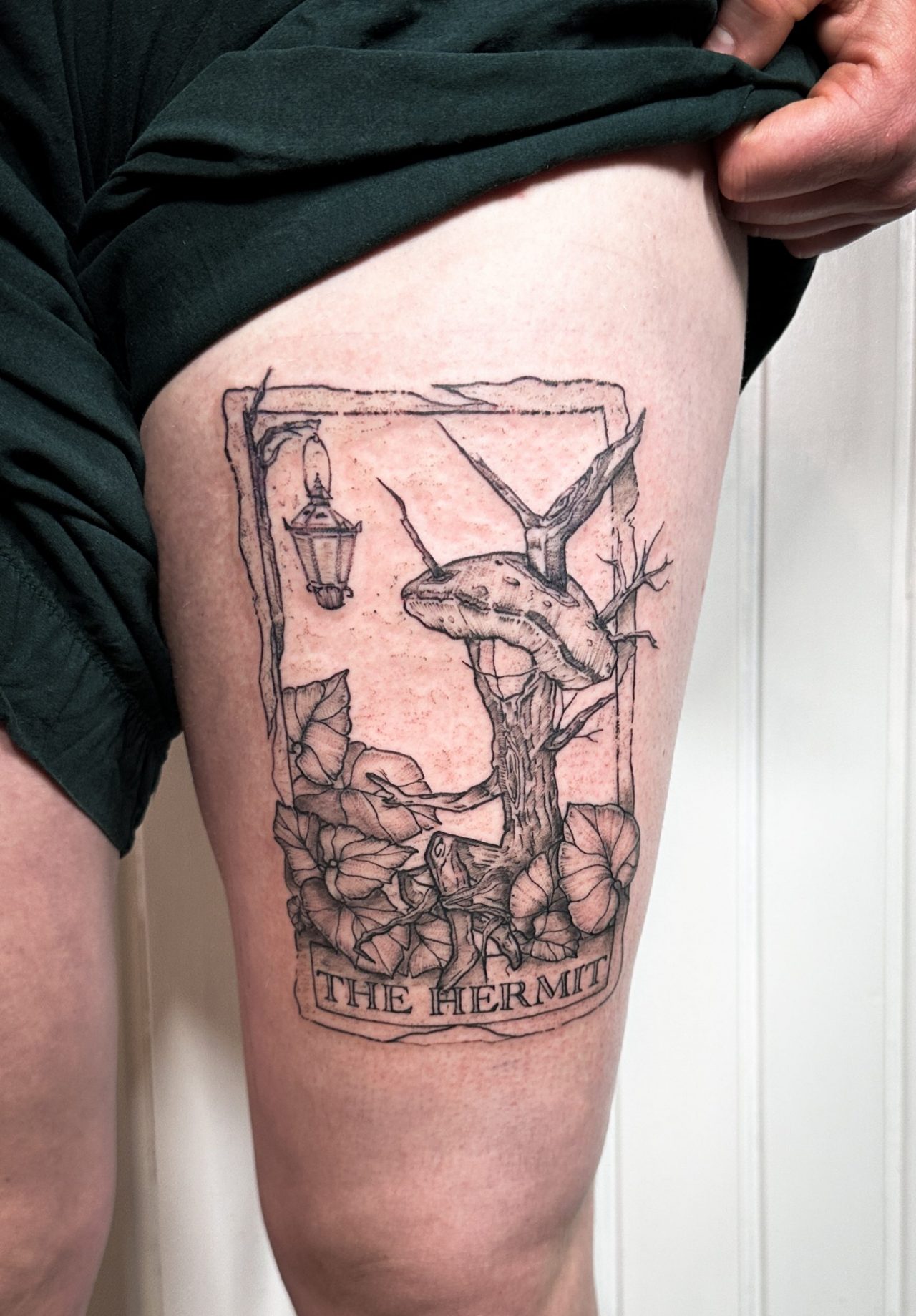 Oberschenkeltattoo: Tarotkarte "The Hermit", Baumfigur mit Pilzkopf sitzt zwischen Blättern mit Laterne